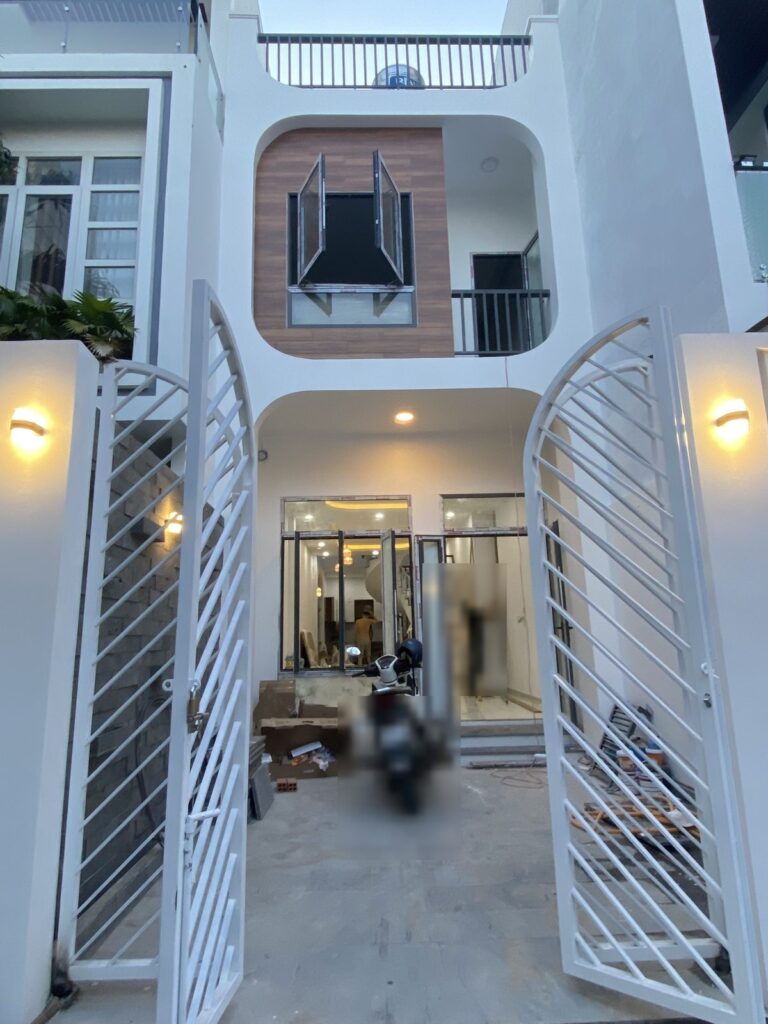 Nhà phố hai tầng cổng sắt vòm trắng, mảng ốp gỗ mặt tiền, đèn đã bật khi bàn giao